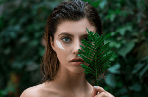 Girl holding leaf to face with moisturizing cream on other side with wet, short hair.